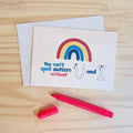 A cream greeting card with a colorful rainbow and the text 'You can't spell autism without U and I' lies on a light wood surface, with a matching white envelope behind it and a bright pink marker in front.
